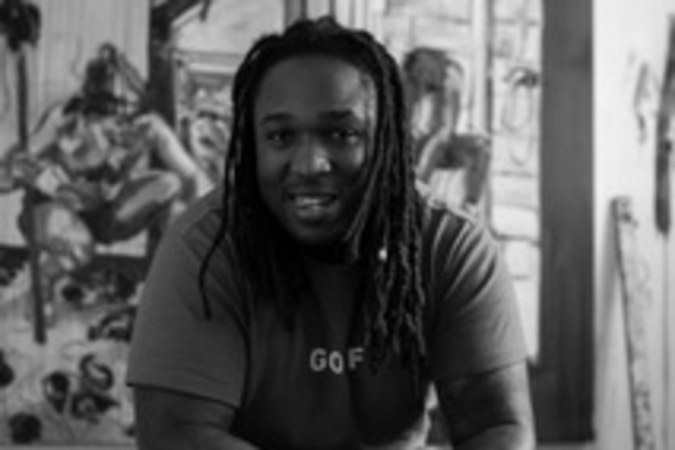 Black and white photograph of the artist Shaquelle Whyte seated, in his studio with his work