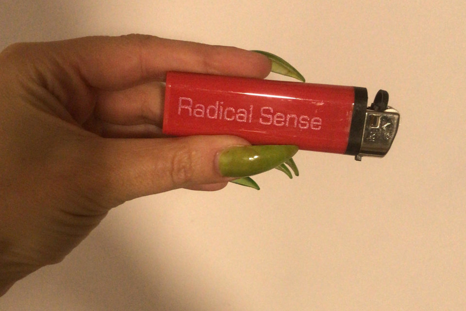 Radical Sense lighter, 2023 A photograph of a thumb and forefinger, with long, varnished transparent lime green nails, holding a red lighter inscribed with the words “Radical Sense” in pink Eurostile type.